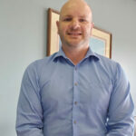Man in blue shirt standing indoors with framed picture behind.