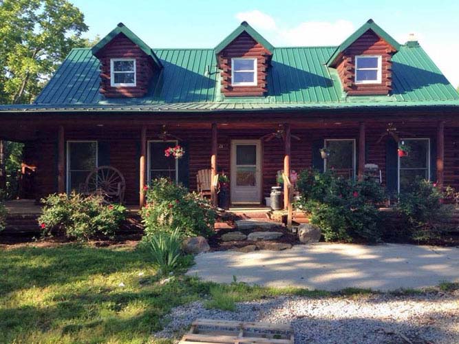Log home with green metal roof, dormers, and front porch.